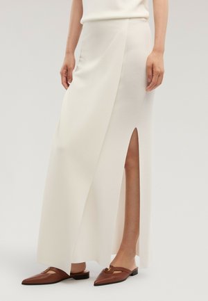 Mujer con falda midi blanca con una abertura frontal y zapatos planos marrones con punta afilada, de pie frente a un fondo liso.