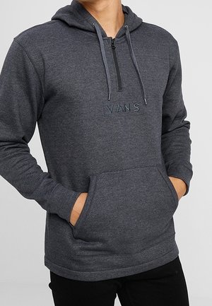 Hombre con sudadera Vans gris oscuro con bolsillo frontal, media cremallera y cordones, manos en los bolsillos, de pie frente a un fondo claro y liso.