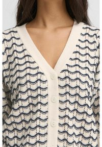 Krämfärgad kofta med marinblå vågmönster, V-ringning och fem knappar. Lätt stickad tyg som skapar en texturerad, öppen design.