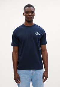 Tamsiai mėlyna medvilninė marškinėliai su trumpomis rankovėmis, apvaliu iškirpimu ir mažu baltu logotipu kairėje krūtinės pusėje. Atrodo puikiai su šviesiai mėlynais džinsais.