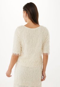 Maglione bianco e morbido con maniche corte, caratterizzato da una vestibilità ampia e una superficie testurizzata con frange. La vista posteriore mette in evidenza lo scollo a barchetta e la morbida caduta.