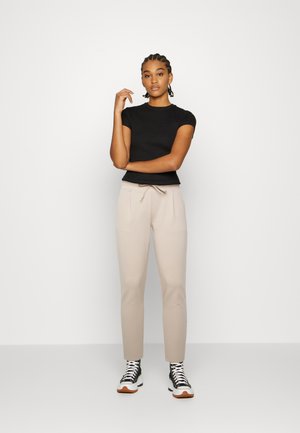 Frau trägt ein schwarzes kurzärmeliges Oberteil, beige Hose mit Kordelzug und schwarz-weiße High-Top-Sneaker, steht vor einem einfachen weißen Hintergrund.