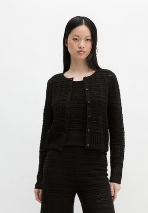 Kobieta o długich czarnych włosach, ubrana w czarny teksturowany sweter na guziki z dzianiny i dopasowane dzianinowe spodnie, stojąca na białym tle.