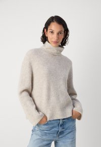 Beige strikket høyhalset genser med rillet tekstur, løs passform og senkede skuldre, kombinert med lyseblå jeans.