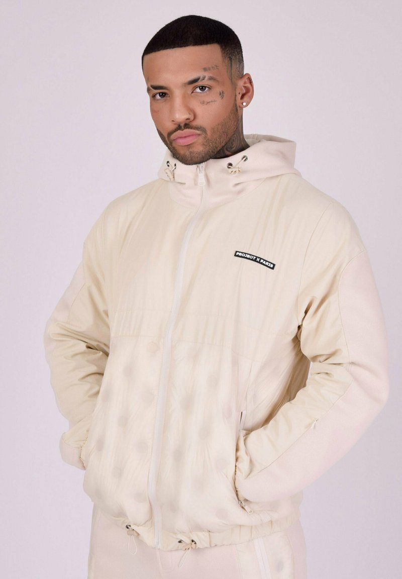 Veste à capuche beige clair avec une texture lisse, dotée d'un zip, d'une taille à cordon de serrage et d'un subtil motif à pois sur la moitié inférieure.