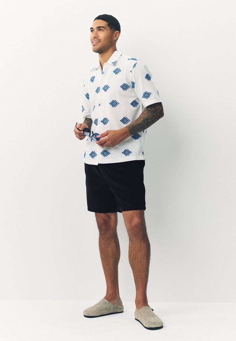 Wit overhemd met korte mouwen en blauw diamantpatroon, zwarte korte broek en grijze instapschoenen. Soepele stof met een ontspannen pasvorm.