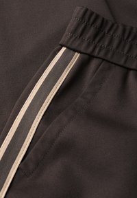 Tessuto marrone scuro con una texture liscia. Presenta una striscia verticale beige e una cintura elastica, enfatizzando un design sportivo.