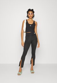 Top corto nero con logo, leggings neri a vita alta con fessure laterali, abbinati a sandali con tacco verde e una borsa bianca.