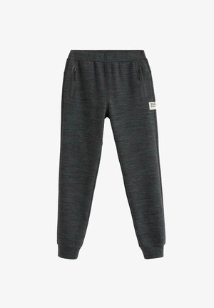 Tienerjongen staand in een casual wit T-shirt, donkere joggingbroek en witte sneakers met blauwe en oranje accenten tegen een effen achtergrond.