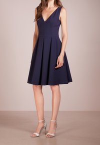 Navyblaues ärmelloses Kleid mit V-Ausschnitt, tailliertem Oberteil und ausgestellt Rock. Kombination mit silbernen Riemchen-Pumps. Glatte Stofftextur.