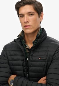 Chaqueta acolchada negra con diseño acolchado y cierre de cremallera. Presenta un cuello alto y el logo de Superdry en el pecho izquierdo. Combinada con una camisa de cuadros verde.
