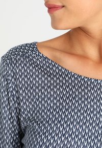 Marinblus med vitt pil-liknande mönster, mjukt tyg, båtformad halsringning och lätt texturerad yta. Fokusera på detaljerna kring halsringningen och mönstret.