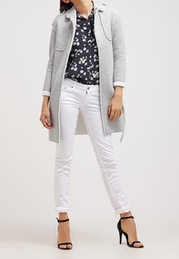 Abrigo largo gris con bolsillos laterales sobre una blusa floral azul marino, combinado con pantalones ajustados blancos y tacones altos negros con tiras.