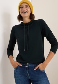 Schwarzer Hoodie mit hohem Kragen und Kordelzügen, kombiniert mit blauen Denim-Jeans. Das Modell trägt eine gelbe gestrickte Mütze.