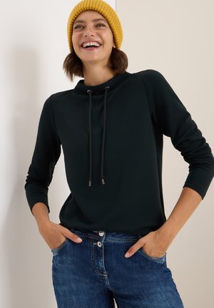 Czarna bluza z wysokim kołnierzem i sznurkiem, zestawiona z niebieskimi dżinsami. Modelka nosi żółtą wełnianą czapkę.