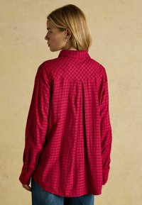 Red plaid button-up shirt with a relaxed fit. Features long sleeves, a collar, and a yoke detail. Made of soft fabric with a textured finish.