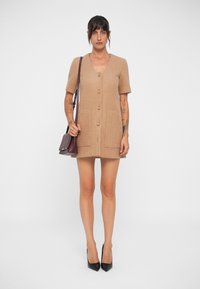 Abito beige corto realizzato in tessuto testurizzato, con scollatura a V, maniche corte, cinque bottoni e due tasche frontali. Tacchi alti neri e una piccola borsa.