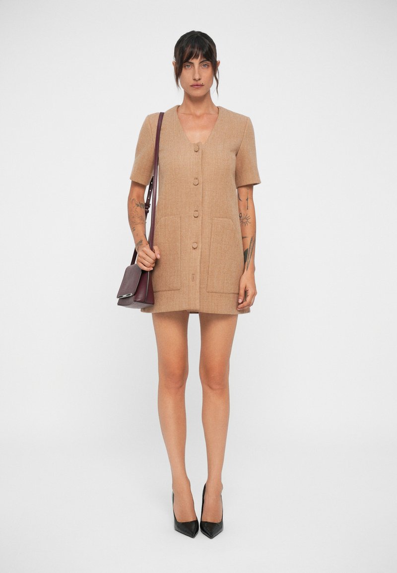 Abito beige corto realizzato in tessuto testurizzato, con scollatura a V, maniche corte, cinque bottoni e due tasche frontali. Tacchi alti neri e una piccola borsa.