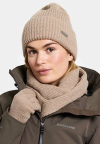 Beige knitted beanie and matching scarf made of textured wool, paired with a brown insulated jacket. Features ribbed pattern and brand label.