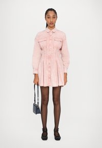 Robe en denim rose clair avec un devant à boutons, deux poches poitrine et une jupe plissée. Modèle tenant un petit sac à main bleu marine, portant des ballerines noires.
