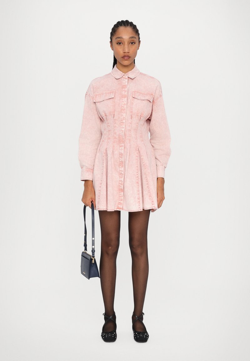 Robe en denim rose clair avec un devant à boutons, deux poches poitrine et une jupe plissée. Modèle tenant un petit sac à main bleu marine, portant des ballerines noires.