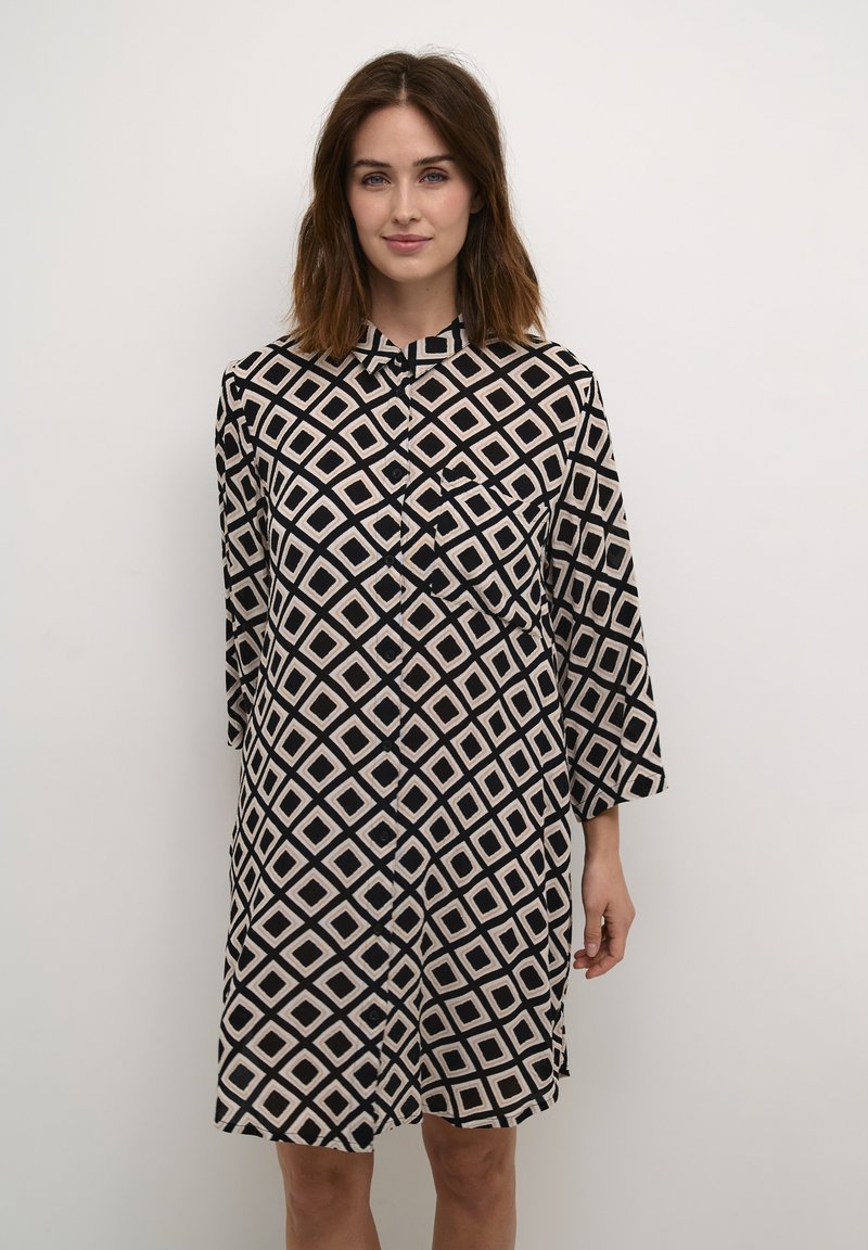 Button-up dress in black and beige with diamond pattern. Features long sleeves, a collar, and a front pocket. Smooth fabric, knee-length.