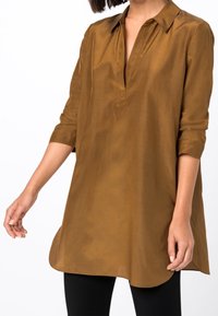 En kvinna klädd i en lång, brun tunika med krage och uppvikta ärmar, tillsammans med svarta leggings, står mot en vit bakgrund.