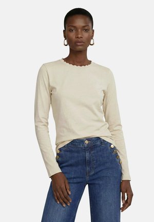 Kvinde med kort hår iført en cremefarvet langærmet top med bølget halsudskæring og højtaljede blå jeans med guldknapper.