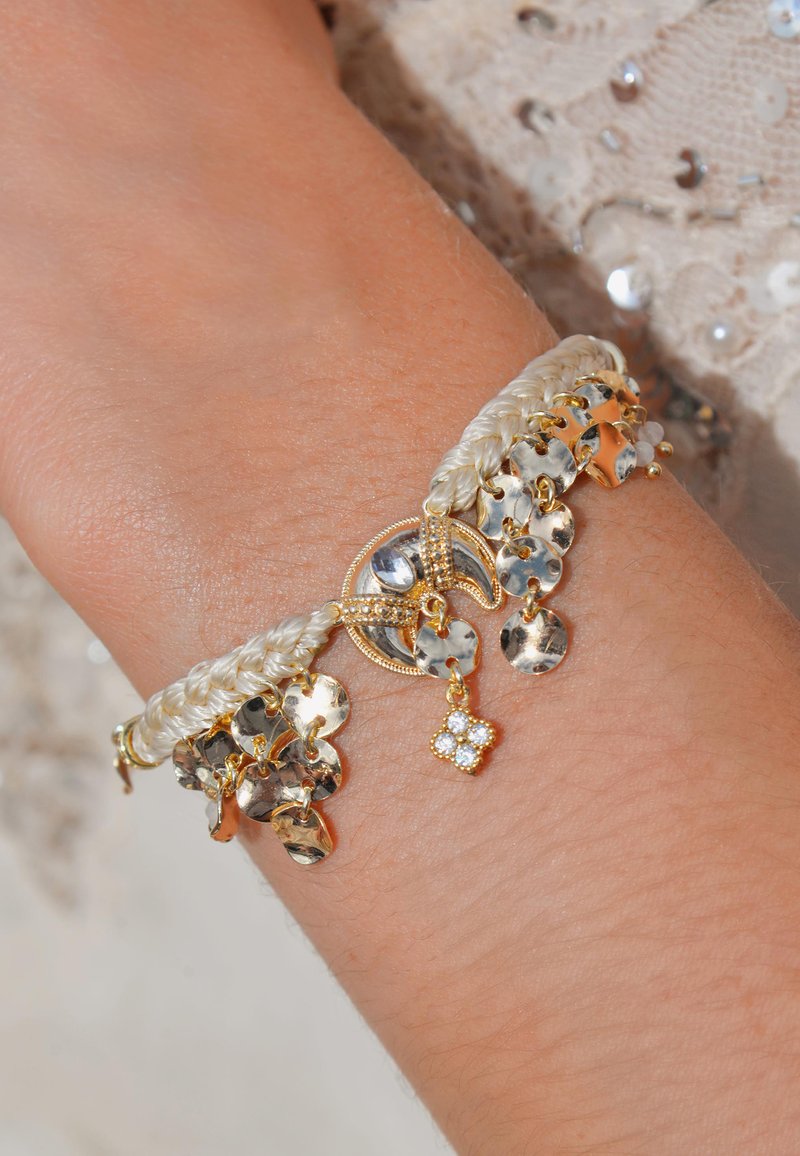 Bracelet doré ornée d'un matériau tressé crème, de disques métalliques placés à la main et d'un petit charm floral décoré de pierres transparentes.