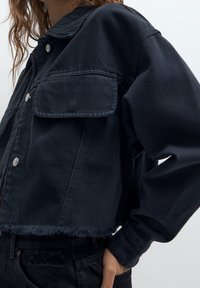 Przycięta czarna kurtka dżinsowa z ostrokątnym kołnierzem, przednią kieszenią i postrzępionym brzegiem. Posiada metalowe zatrzaski oraz długie, luźne rękawy.