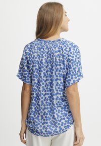 Blusa blu e bianca con una vestibilità ampia, maniche corte e un collo arricciato. Presenta un motivo a pois di varie dimensioni.