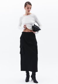Zwarte high-waisted rok met twee frontzakken, gecombineerd met een witte top met lange mouwen en zwarte hakschoenen, en een tas met textuur.