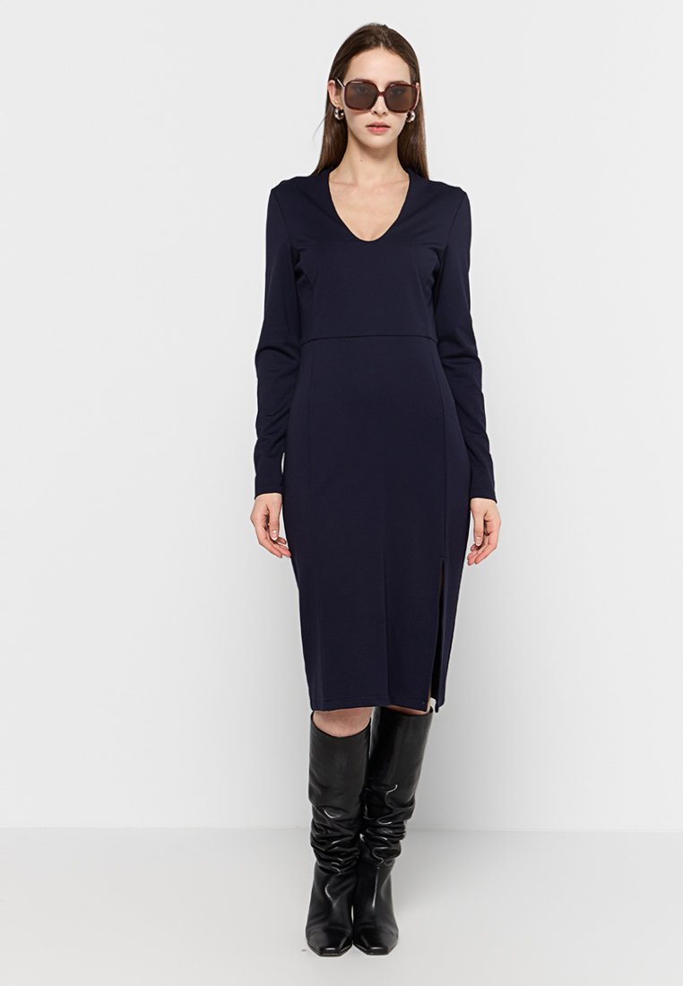 Abito lungo blu navy con maniche lunghe, scollatura a V e spacco laterale, realizzato in un tessuto morbido. Abbinato a stivali neri alti fino al ginocchio e occhiali da sole.