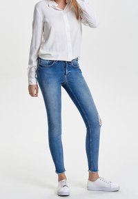 Jeans ajustados azules con un desgaste sutil y adornos rasgados en las rodillas, combinados con una camisa blanca de botones y zapatillas blancas.