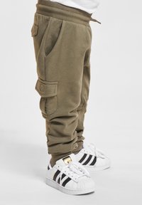 Ett barn i olivgröna cargojoggers och vita Adidas-sneakers med svarta ränder, stående mot en vit bakgrund.