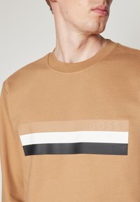 Felpa marrone a maniche lunghe con logo testurizzato e design a blocchi orizzontali in beige, bianco e nero sulla parte superiore.