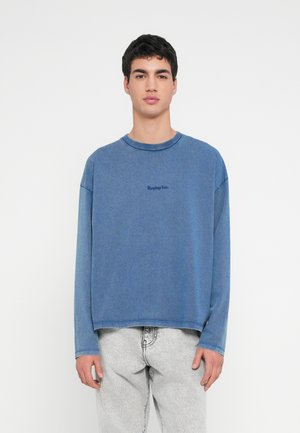 Giovane uomo con capelli scuri e corti indossa una camicia blu a maniche lunghe larga con la scritta "Replay Inc." e jeans grigi chiaro, in piedi contro uno sfondo bianco.