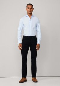 Muškarac u svijetloplavoj košulji na dugme, crnim hlačama i smeškim suéde cipelama stoji naslonjen na jednostavnu svijetlosivu pozadinu.