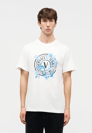 Jonge man die een wit Versace Jeans Couture T-shirt draagt met een blauw swirl-ontwerp en zwarte broek, staand tegen een effen achtergrond.
