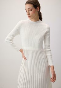 Vrouw met bruin haar in een lage paardenstaart draagt een witte geribbelde jurk met lange mouwen, staat met één hand op de heup en kijkt omlaag naar de zijkant.