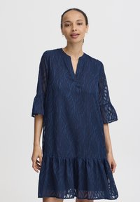 Rochie bleumarin cu margine învolutată în straturi, având un model texturat în valuri, decolteu în V și mâneci tip clopot. Material ușor, semi-transparent.