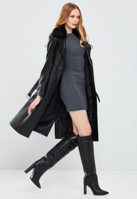 Cappotto di pelle nera con colletto in pelliccia, abito grigio aderente e stivali neri con tacco alti fino al ginocchio. Texture liscia e design snodato.