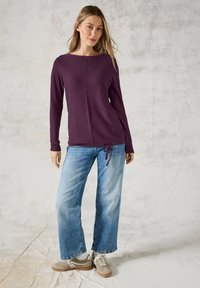 Lila Langarmshirt mit Bootsausschnitt, strukturiertem Stoff und einem Kordelzug in der Taille, kombiniert mit weit geschnittenen hellblauen Jeans.