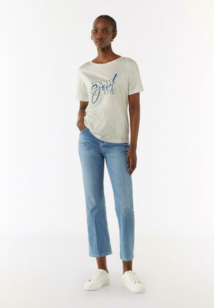 T-shirt gris clair avec lettrage bleu, coupe décontractée, associé à un jean bleu taille mi-haute et des baskets blanches. Tissu lisse et design droit.