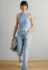 Helle blaue, gerippte Tanktop, mittelblaues, gerades Jeans mit Patches und cremefarbene Absätze. Graue gemusterte Jacke über dem Arm drapiert.