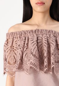 Top off-shoulder in morbido mauve con intricate rifiniture in pizzo. Presenta un bordo arricciato e margini scallopati con piccoli dettagli in perline.
