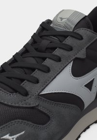 Κλείσιμο εικόνας μαύρου και γκρι αθλητικού παπουτσιού με μαύρα κορδόνια και λευκό λογότυπο Mizuno στο πλάι και στη γλώσσα.