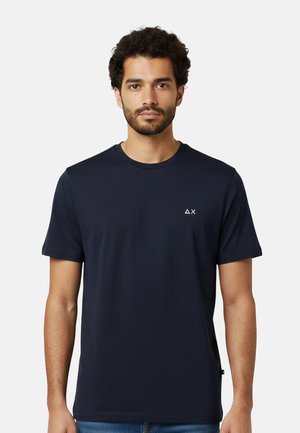 Uomo con capelli ricci e barba che indossa una semplice maglietta blu scuro con un piccolo triangolo bianco e il logo "X" sul petto a sinistra.