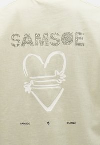 Hellbeiges T-Shirt mit dem Text "SAM"SØ" in verschiedenen Schriftarten und einem weißen Herzgrafik darunter zentriert, mit schwarzem Markenlogo unten.