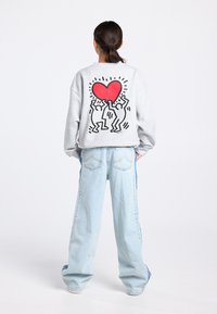 Sweat-shirt gris avec un grand cœur rouge et deux silhouettes dansantes imprimées dans le dos, associé à un jean bleu clair à jambes larges.